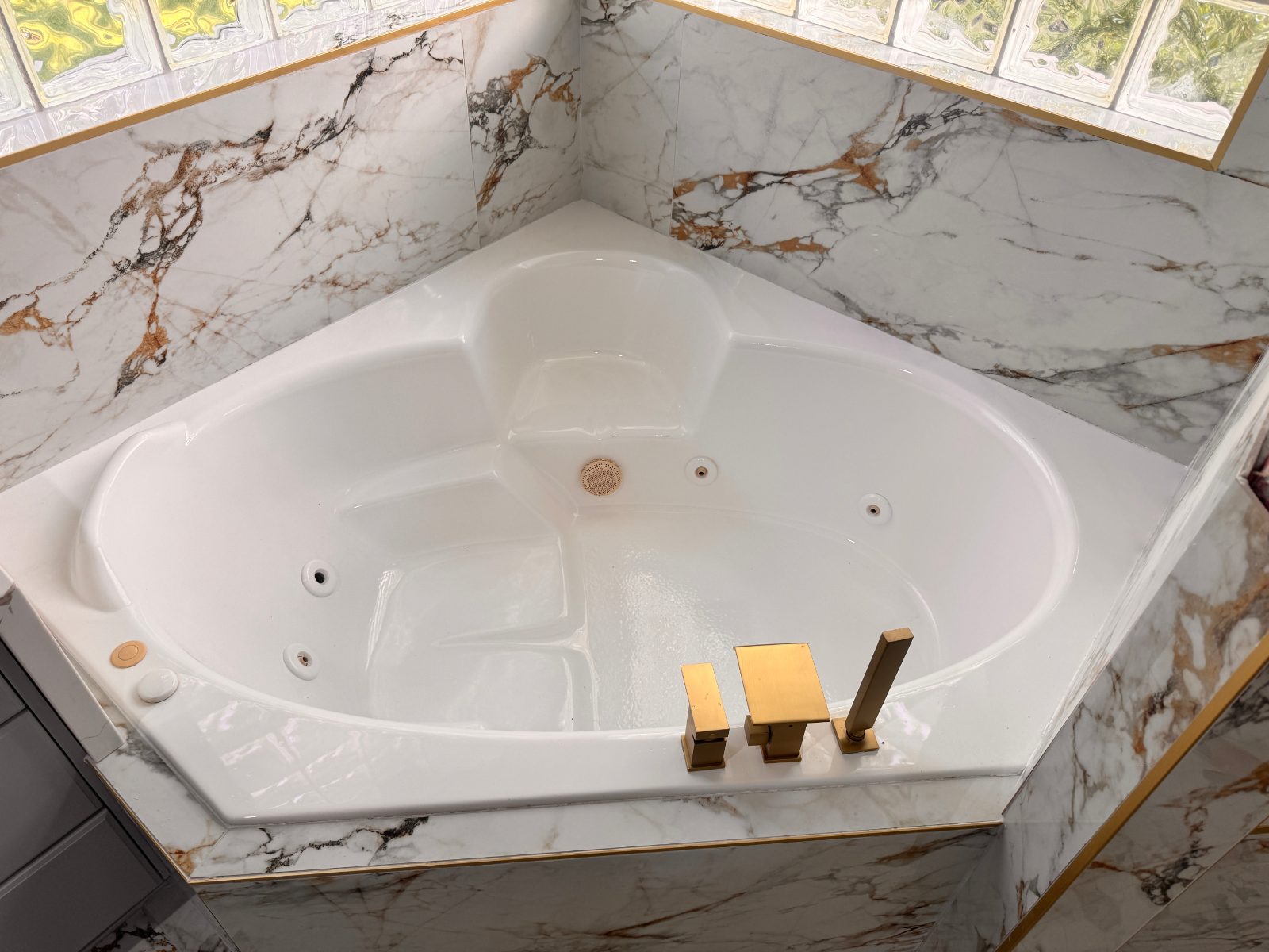 A corner whirlpool spa tub refinished to a bright high-gloss white, framed by a white-and-gold marble surround and glass-block window